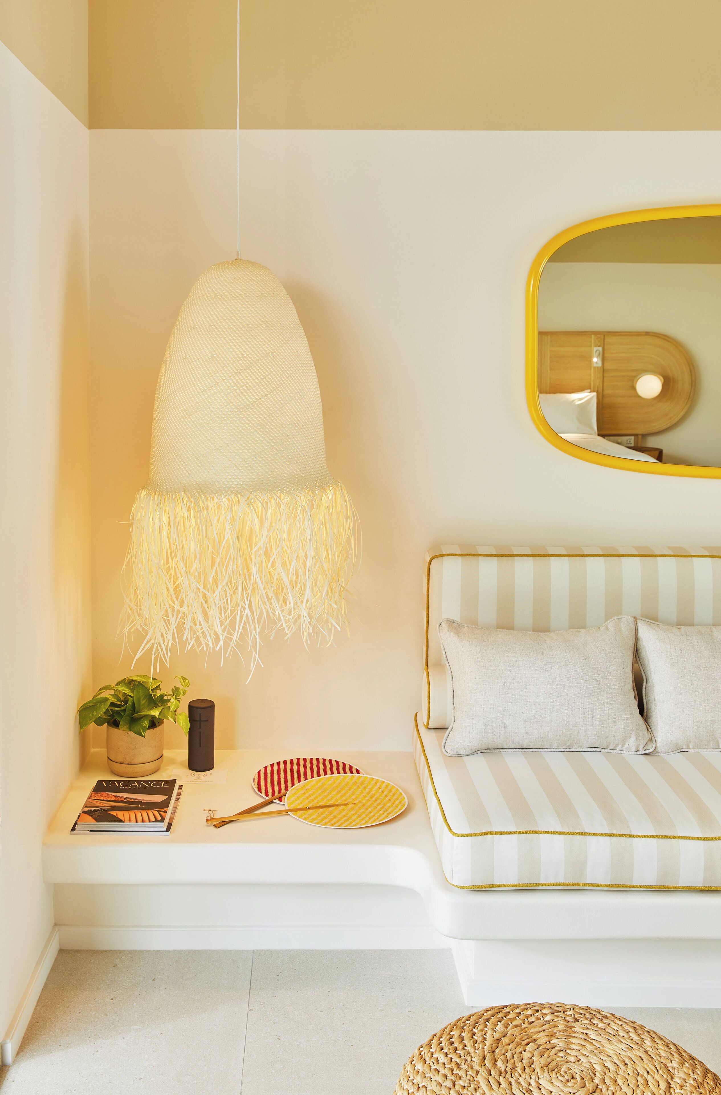 Un dormitorio moderno con un sofá blanco, una lámpara colgante, un espejo y una mesita de noche con un libro y un objeto decorativo.