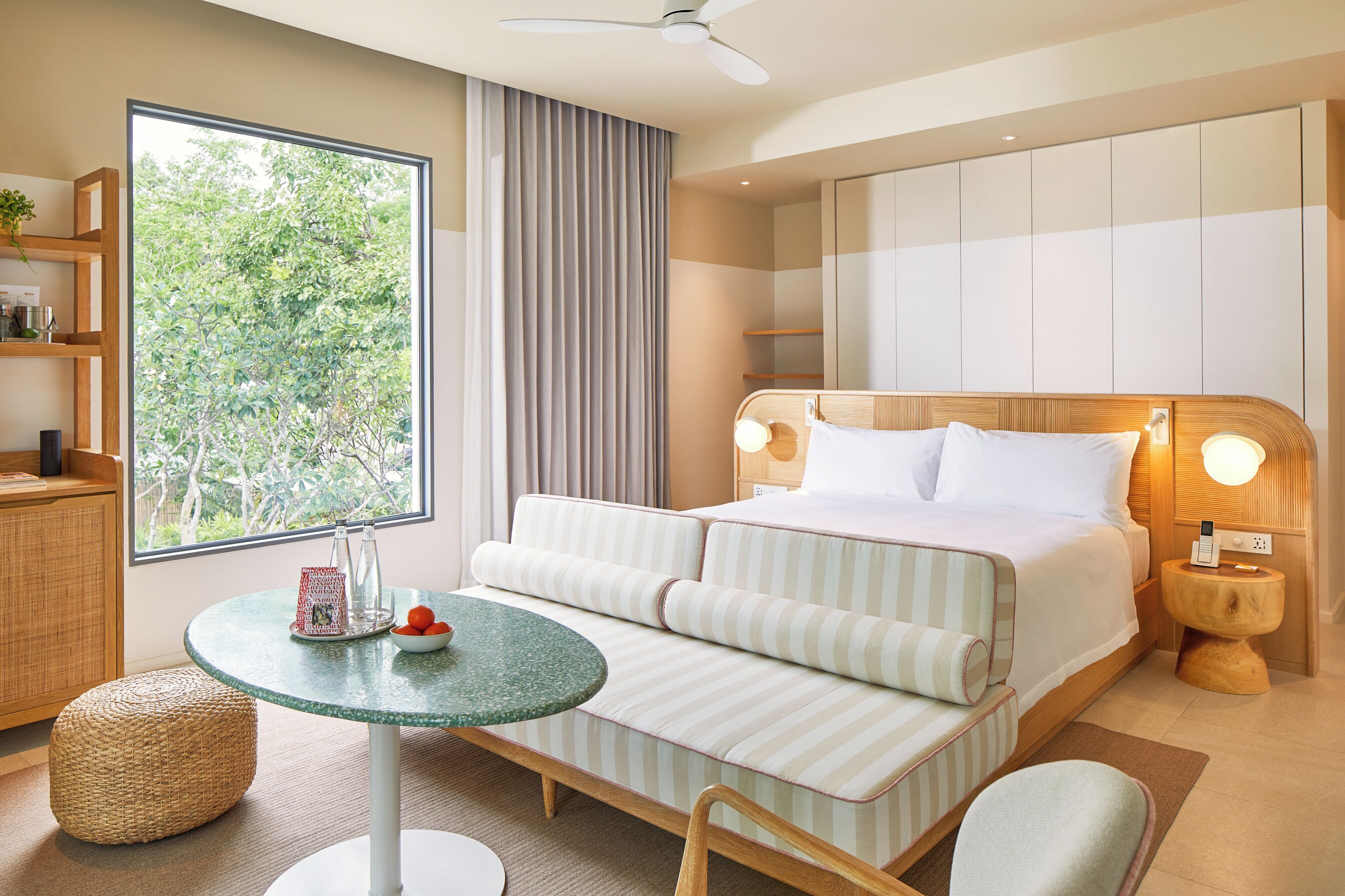 A hotel room with a bed, a round table, a chair, a small stool, a bedside table with a lamp, a shelf with a plant, and a large window with curtains.