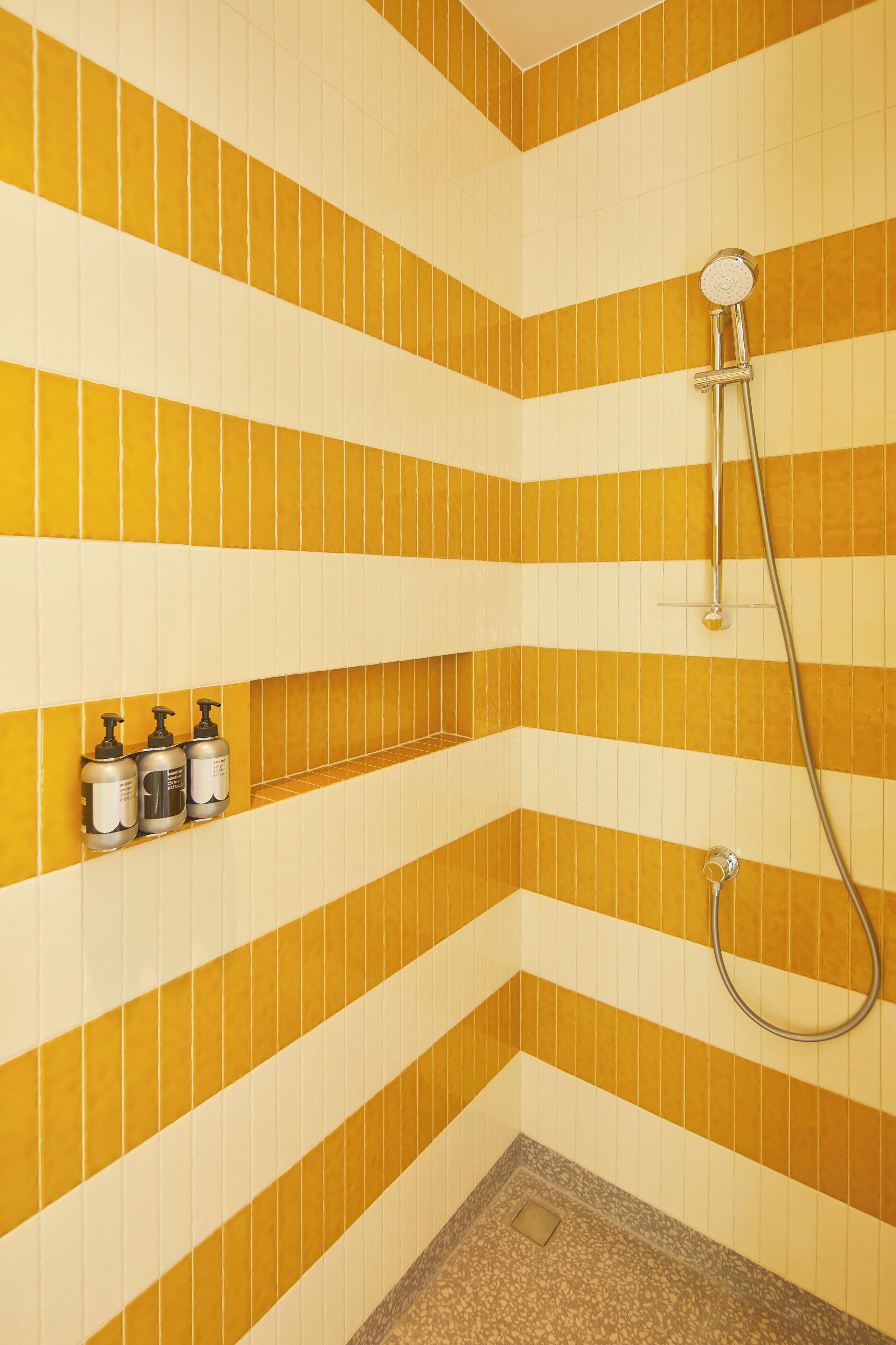 A bathroom with yellow and white striped tiles, a shower, and three bottles on the wall.