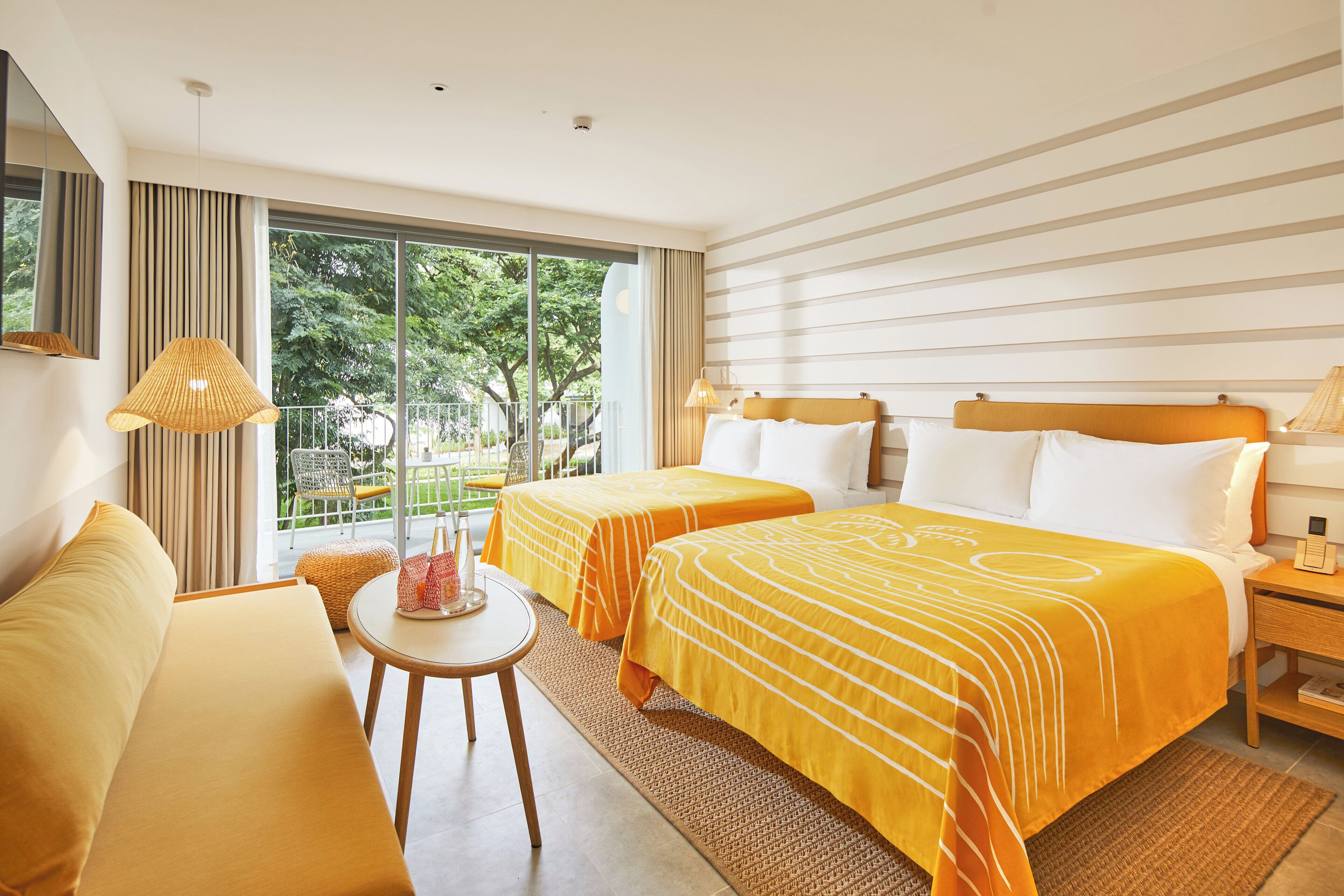 Een hotelkamer met twee bedden, een sofa, een kleine tafel en een balkon met uitzicht op groen.