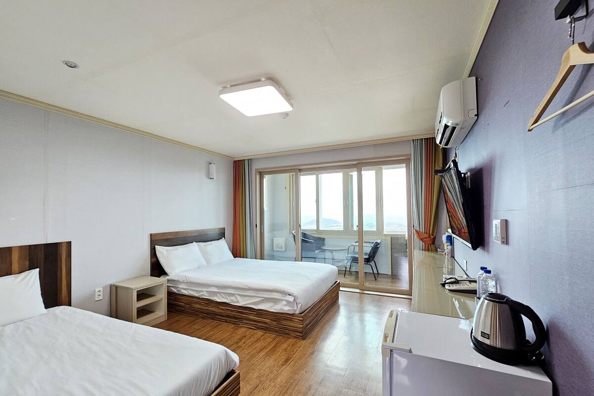 A hotel room with two beds, a desk, a refrigerator, a kettle, and a sliding glass door leading to a balcony with a view.