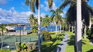 Una cancha de tenis con un camino, palmeras y vista al mar con barcos.