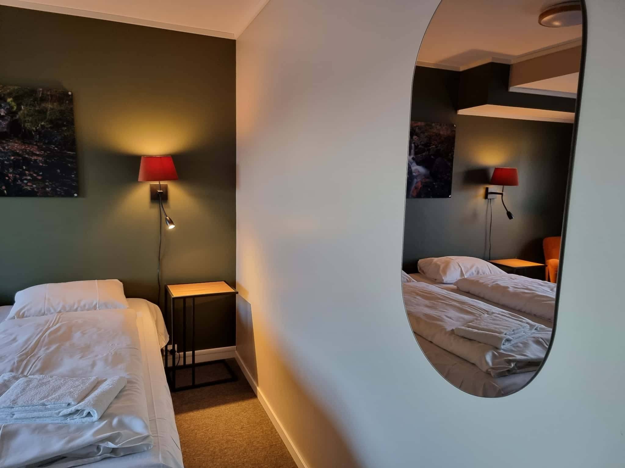 A hotel room with two beds, a small table, a mirror, and two wall-mounted lamps.