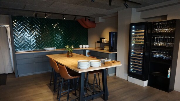 A modern kitchen with a wooden table, bar stools, a herringbone-patterned backsplash, and a wine refrigerator. - Verdde Hotel Lakselv (Porsanger)
