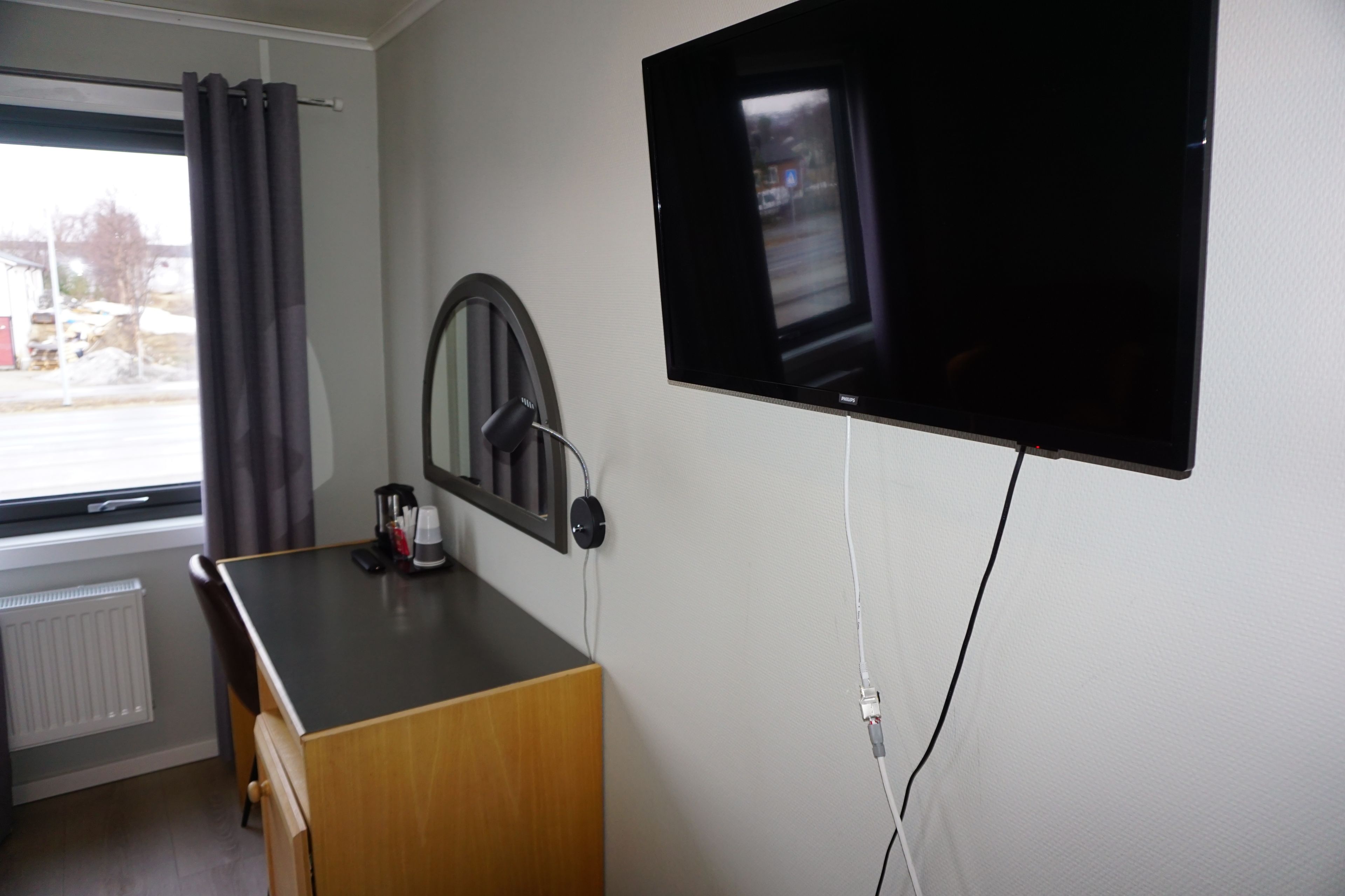A room with a television mounted on the wall, a desk with a chair, a window with curtains, and a radiator.