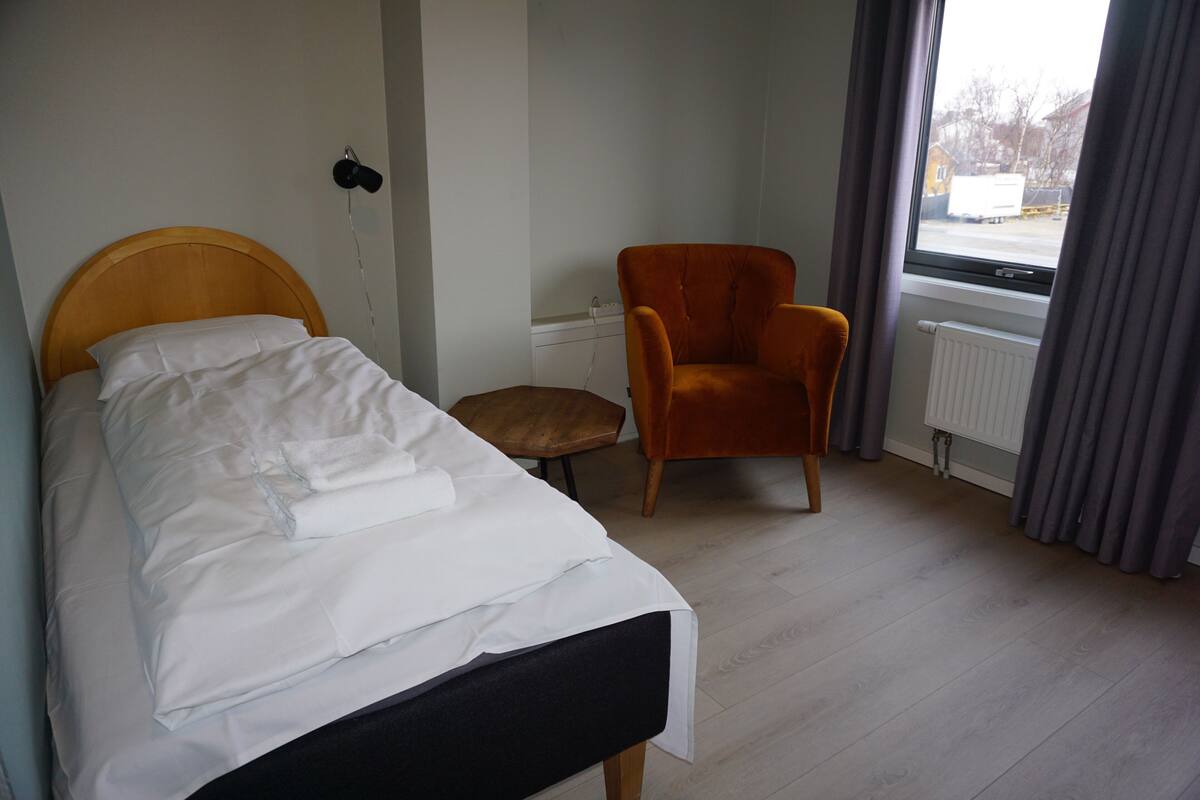 A bedroom with a bed, a chair, a small stool, a window, and a radiator.