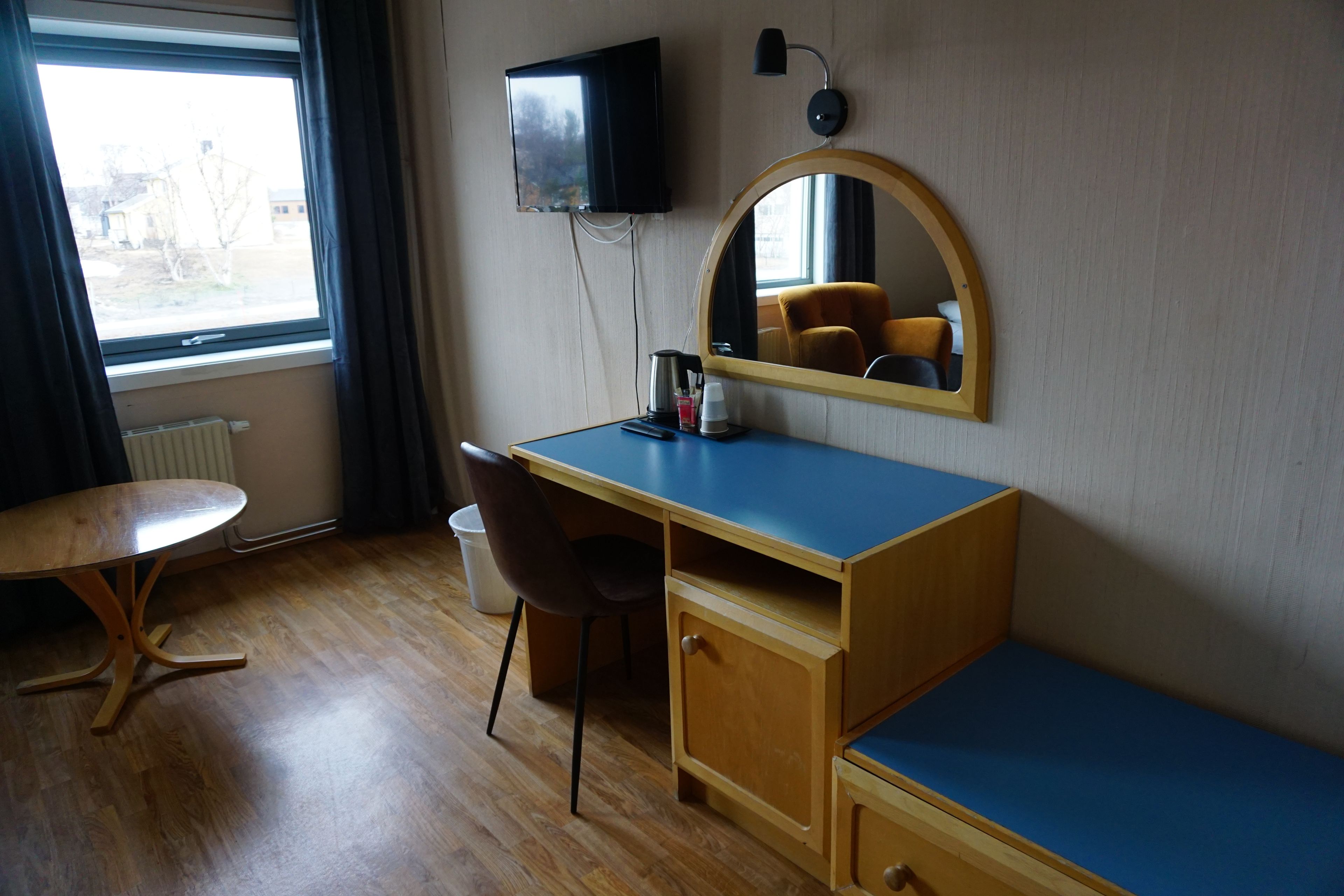 A room with a wooden desk, a chair, a television mounted on the wall, and a window with curtains.