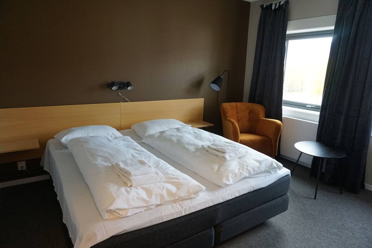 A hotel room with a double bed, a small round table, an orange armchair, and a window with curtains.