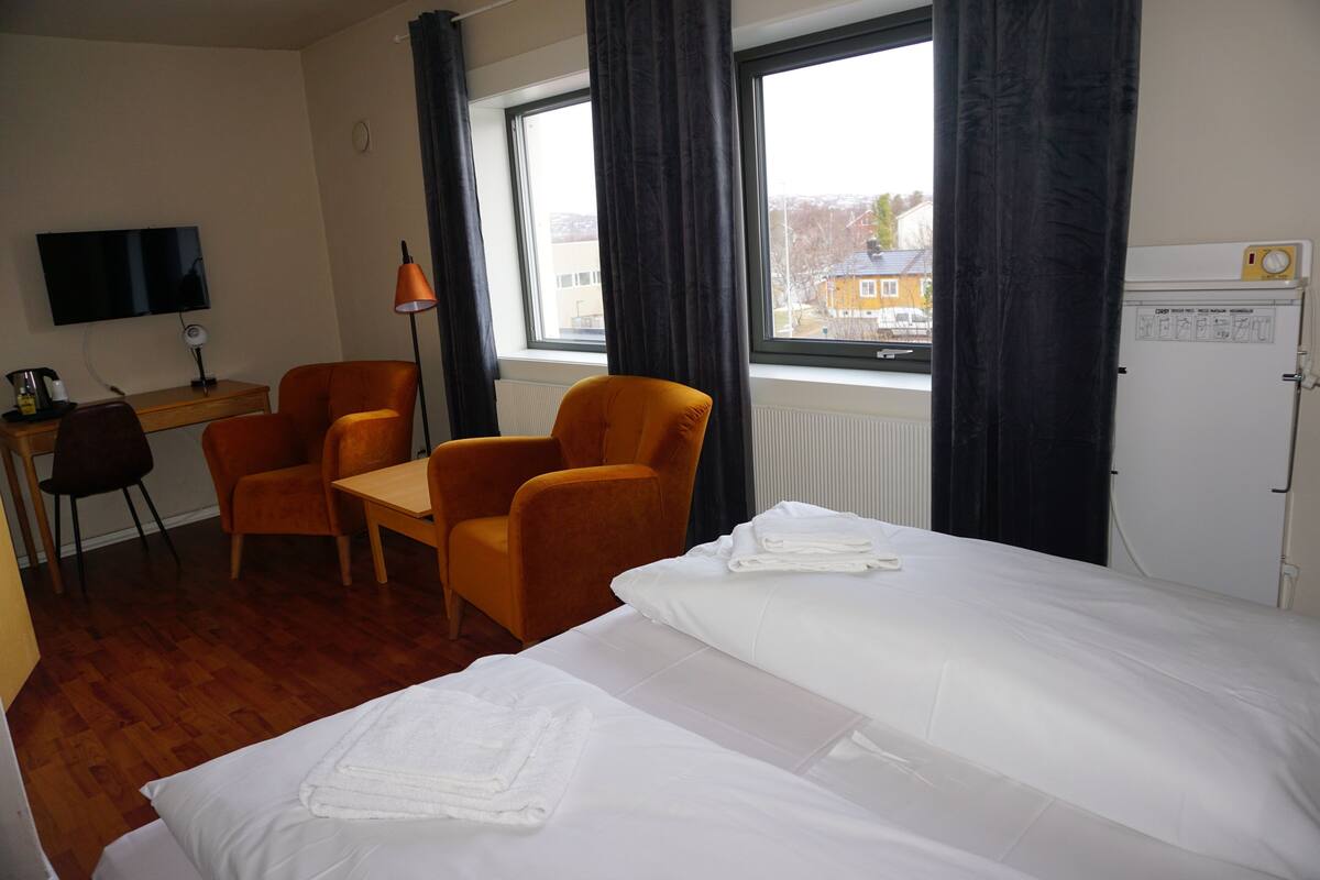 A hotel room with a bed, two orange chairs, a desk, a television, and a window with curtains.