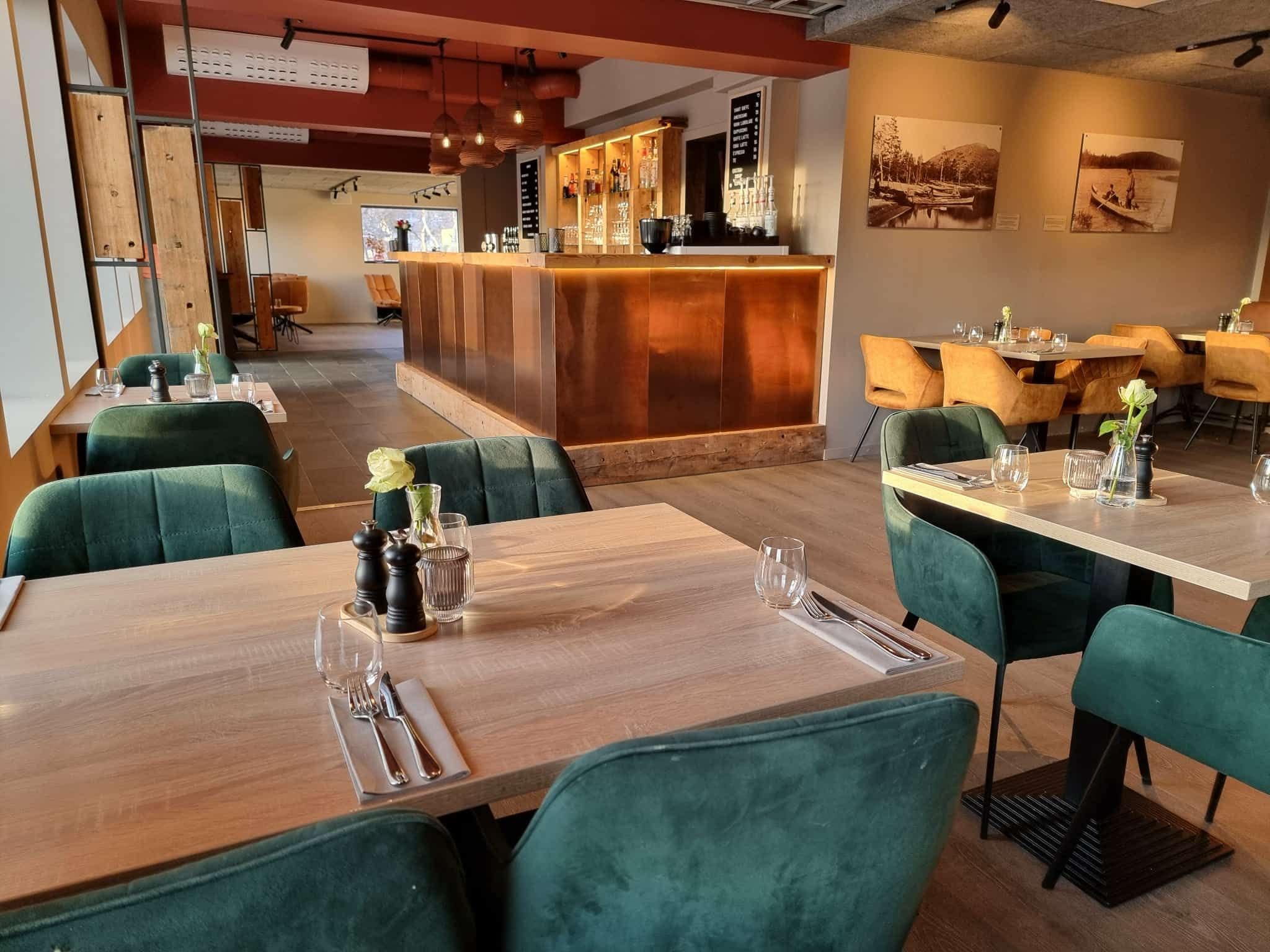 A restaurant interior with wooden tables, green upholstered chairs, a bar area, and wall-mounted artwork.