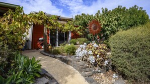 A garden with a variety of plants, a large sunflower sculpture, and a house with a red door and a garage. - Fleurieu Eco Escape - Luxurious, Stylish, Cosy and Clean Hideaway (Aldinga)