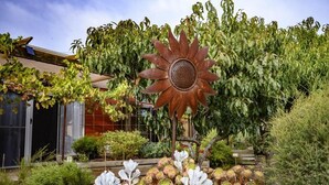 A garden with a large rust-colored sunflower sculpture, various plants, and a building with a red facade in the background. - Fleurieu Eco Escape - Luxurious, Stylish, Cosy and Clean Hideaway (Aldinga)