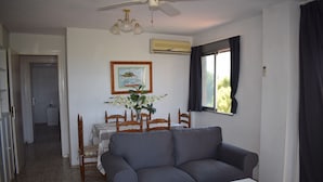 Ein Wohnzimmer mit grauem Sofa, weißem Tisch und einem Holzesstisch mit Stühlen. Ein Deckenventilator, eine Klimaanlage und ein Fenster mit Vorhängen sind vorhanden.