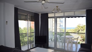 Ein Wohnzimmer mit einem Flachbildfernseher, einem weißen Couchtisch und einer Couch. Es gibt einen Balkon mit Tisch und Stühlen und Blick auf das Meer und die Gebäude.