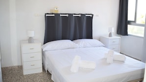 Ein ordentlich bezogenes Bett mit weißen Leinentüchern und aufgerollten Handtüchern, ein Nachttisch mit Lampe und ein Fenster mit dunklen Vorhängen.