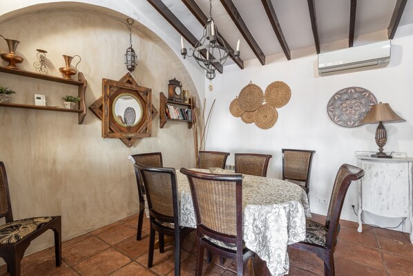 A dining area with a table set for six, surrounded by wicker chairs, a chandelier, and wall decorations including a mirror and woven wall hangings. - Villa "madre tierra pool jacuzzi" with Mountain View, Pool, Whirlpool (Cómpeta)
