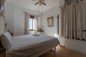 Une chambre à coucher comprenant un lit, un ventilateur de plafond, une fenêtre avec des rideaux, une commode et un miroir.