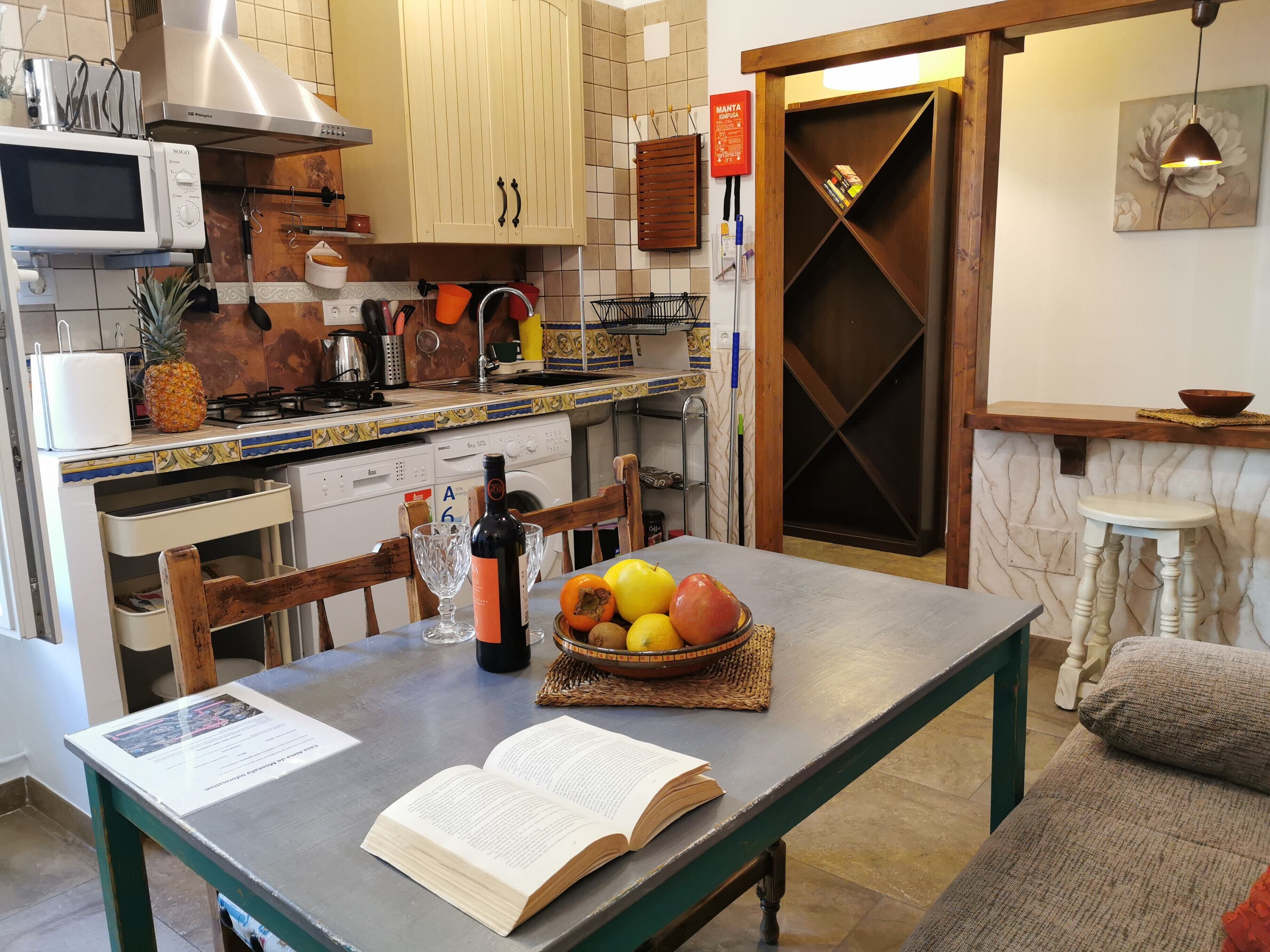 A kitchen with a microwave, stove, and wooden cabinets. There's a dining table with a book and fruit bowl.