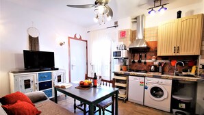 Een ruimtelijke leefruimte met een bank, televisie, eettafel, keuken inclusief fornuis en wasmachine, en een plafondventilator.