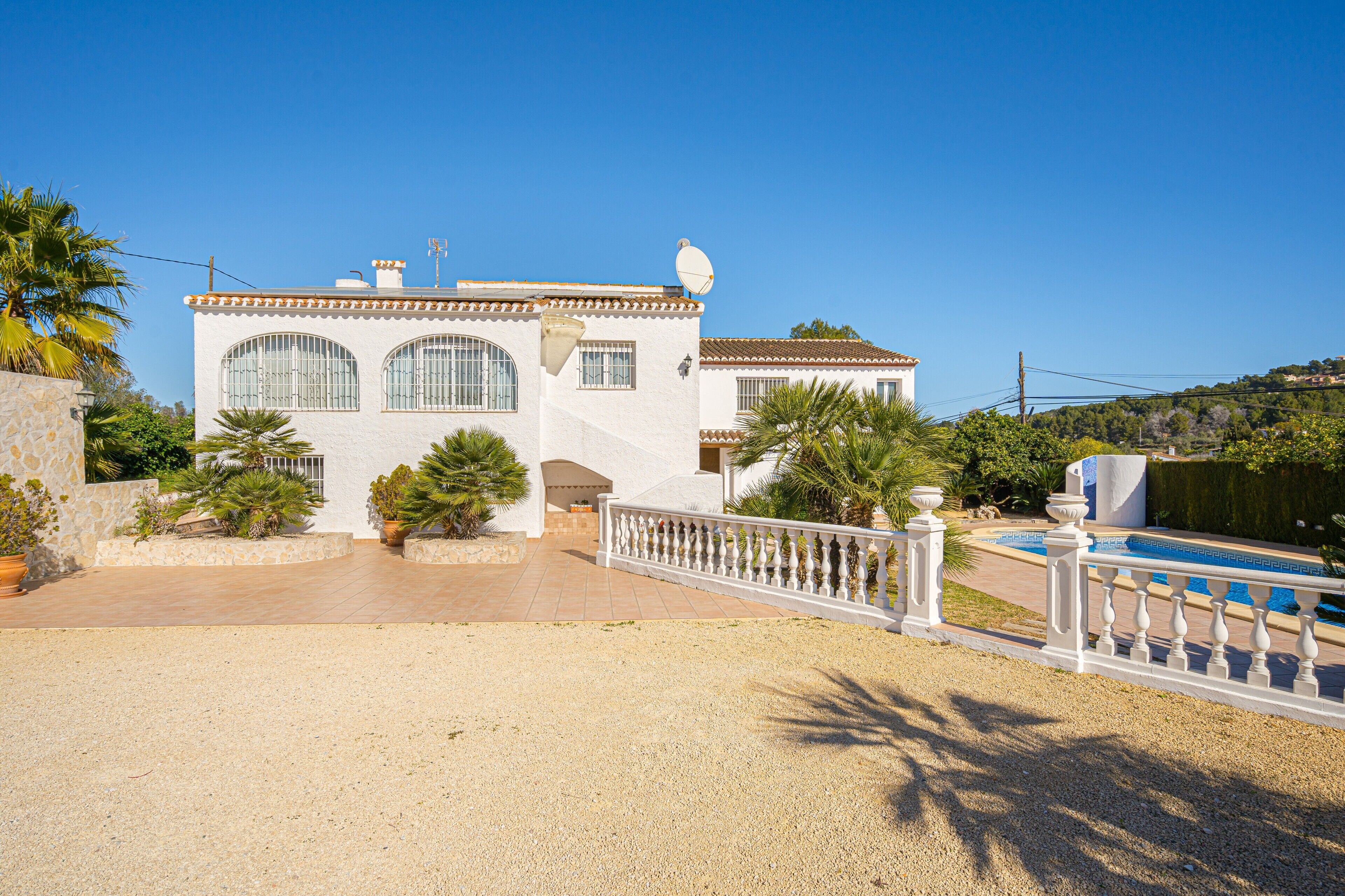 A white villa with a swimming pool, a gravel driveway, and a white fence.