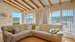Un canapé d’angle beige agrémenté de coussins à motifs floraux, des poutres en bois au plafond et de grandes fenêtres avec des rideaux.