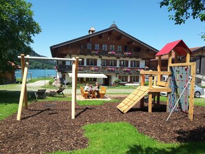 Ein Spielplatz mit Schaukeln und Klettergerüsten, einem Tisch mit Stühlen sowie ein Gebäude im Hintergrund mit Balkonen und Blumenkästen.