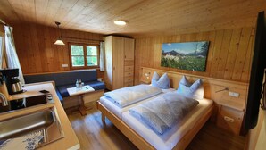 A wooden cabin bedroom with a bed, a bench, a table with a kettle, and a painting on the wall.