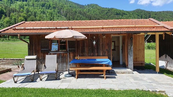 A rustic wooden cabin with a tiled roof, a bench, a table with a book, and a small outdoor kitchen area.