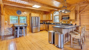 Une cuisine rustique, située dans une cabane en bois, avec un îlot central, des appareils en acier inoxydable et une grande fenêtre.