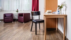 Een kamer met een houten vloer, twee paarse fauteuils, een zwarte stoel, een houten bureau met een bloevas en een rood gordijn.