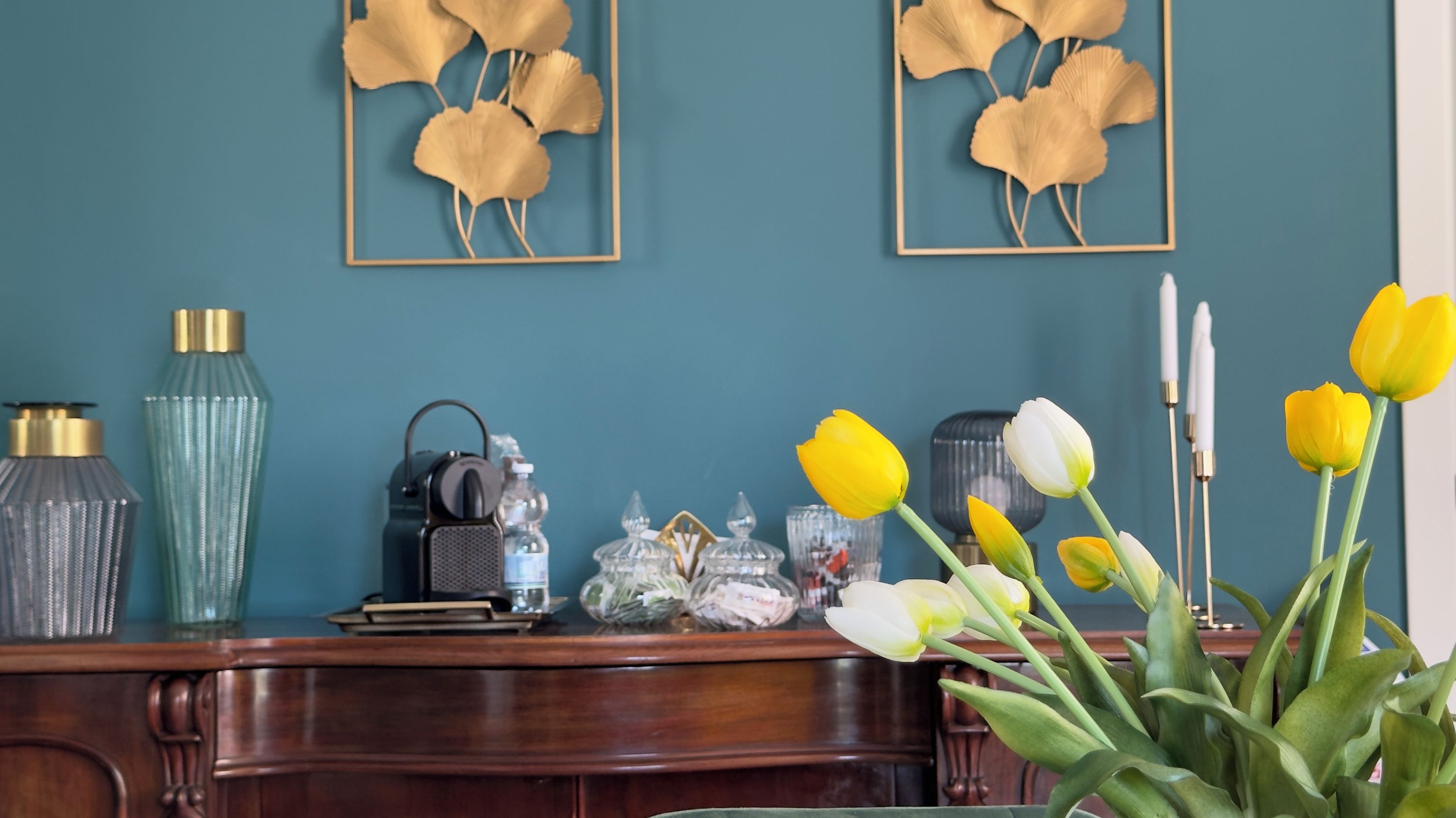 Ein Schreibtisch mit Dekoration, eine Vase mit Blumen und gerahmte Kunstwerke an einer türkisfarbenen Wand.
