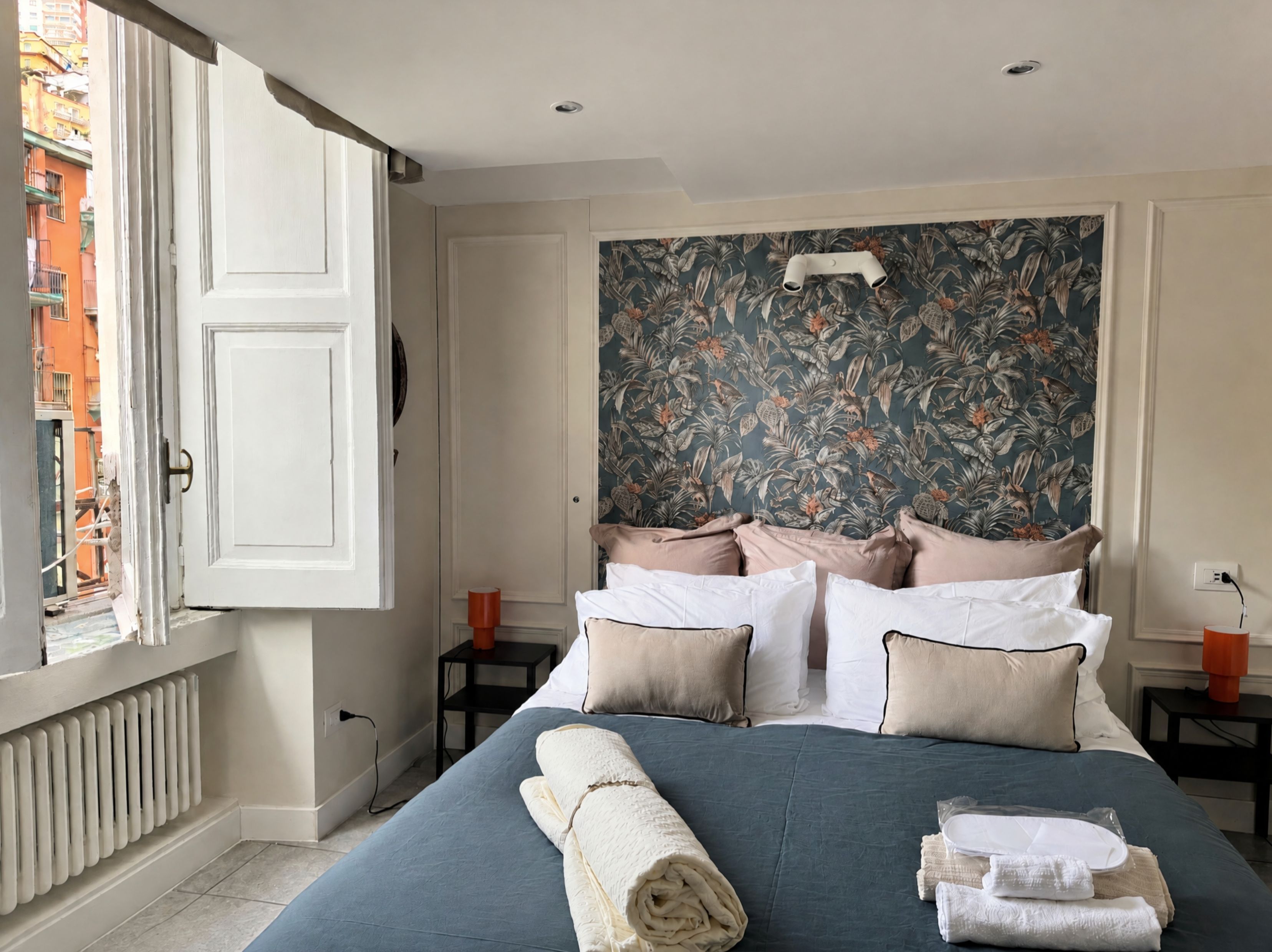 A hotel room with a bed, a window with an open view, a headboard with a floral wallpaper, and a bedside table with a lamp.