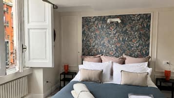 A hotel room with a bed, a window with an open view, a headboard with a floral wallpaper, and a bedside table with a lamp.