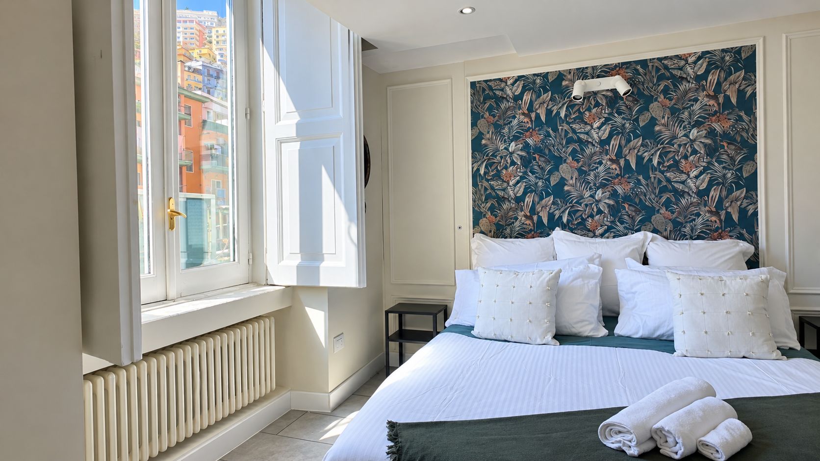 Une chambre d’hôtel avec un grand lit, une fenêtre avec des stores, un radiateur et un papier peint à motifs floraux.