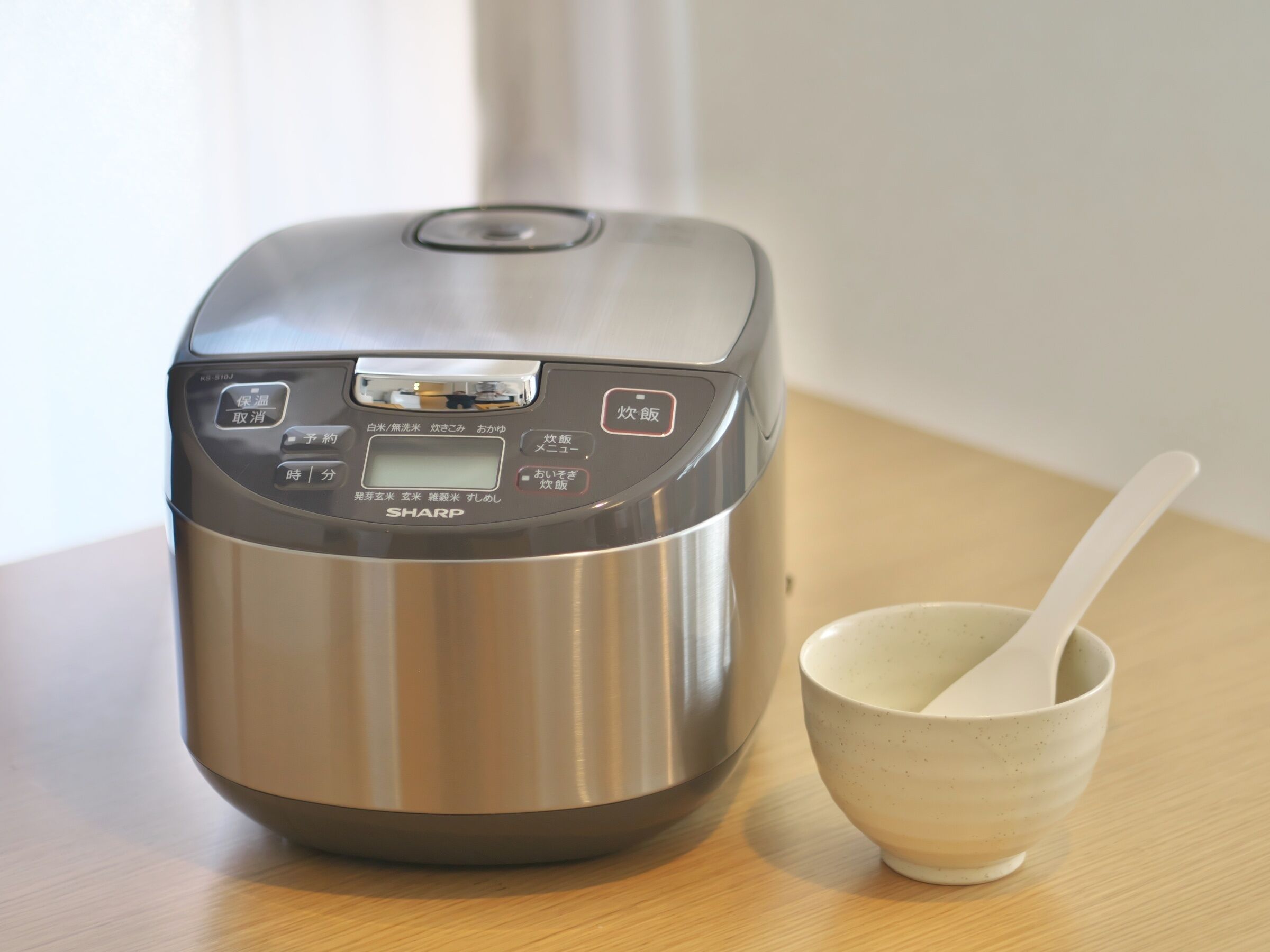 A stainless steel rice cooker with a digital display and control buttons, placed on a wooden surface next to a ceramic bowl with a spoon.