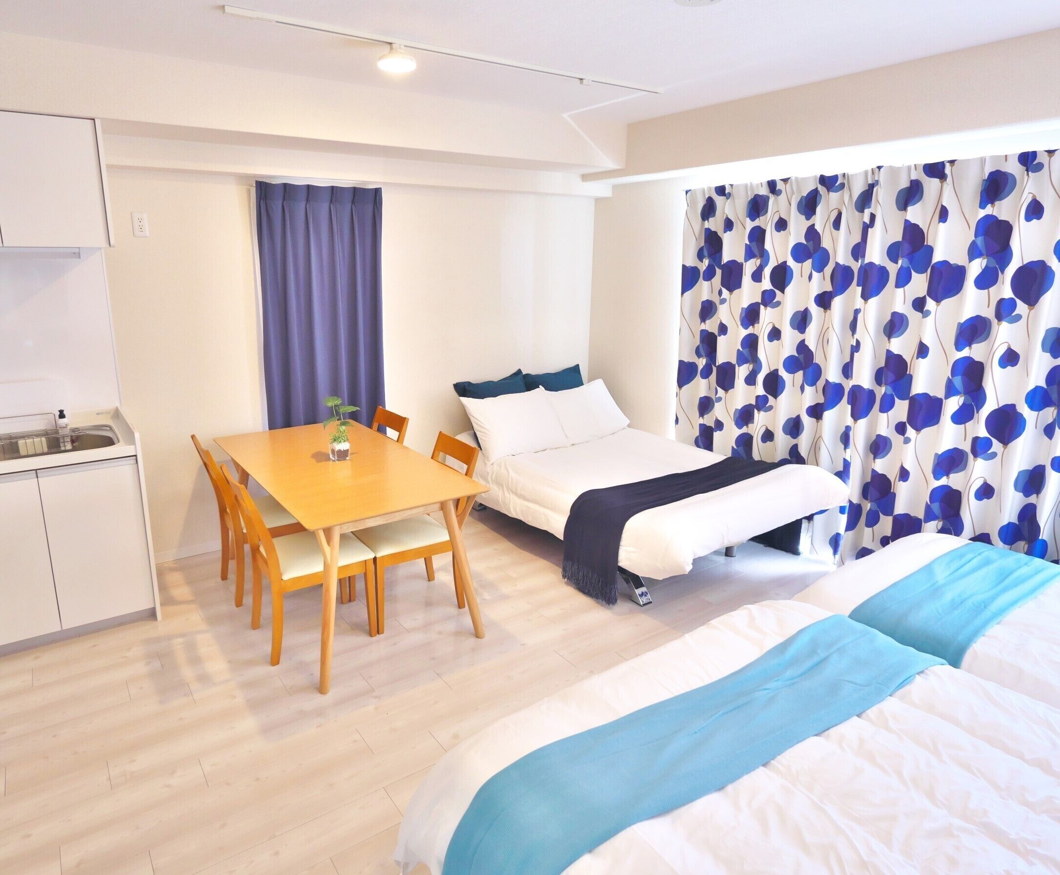 A compact hotel room with a bed, a small table with chairs, a kitchenette, and a window with curtains.