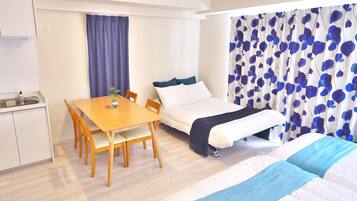 A compact hotel room with a bed, a small table with chairs, a kitchenette, and a window with curtains.