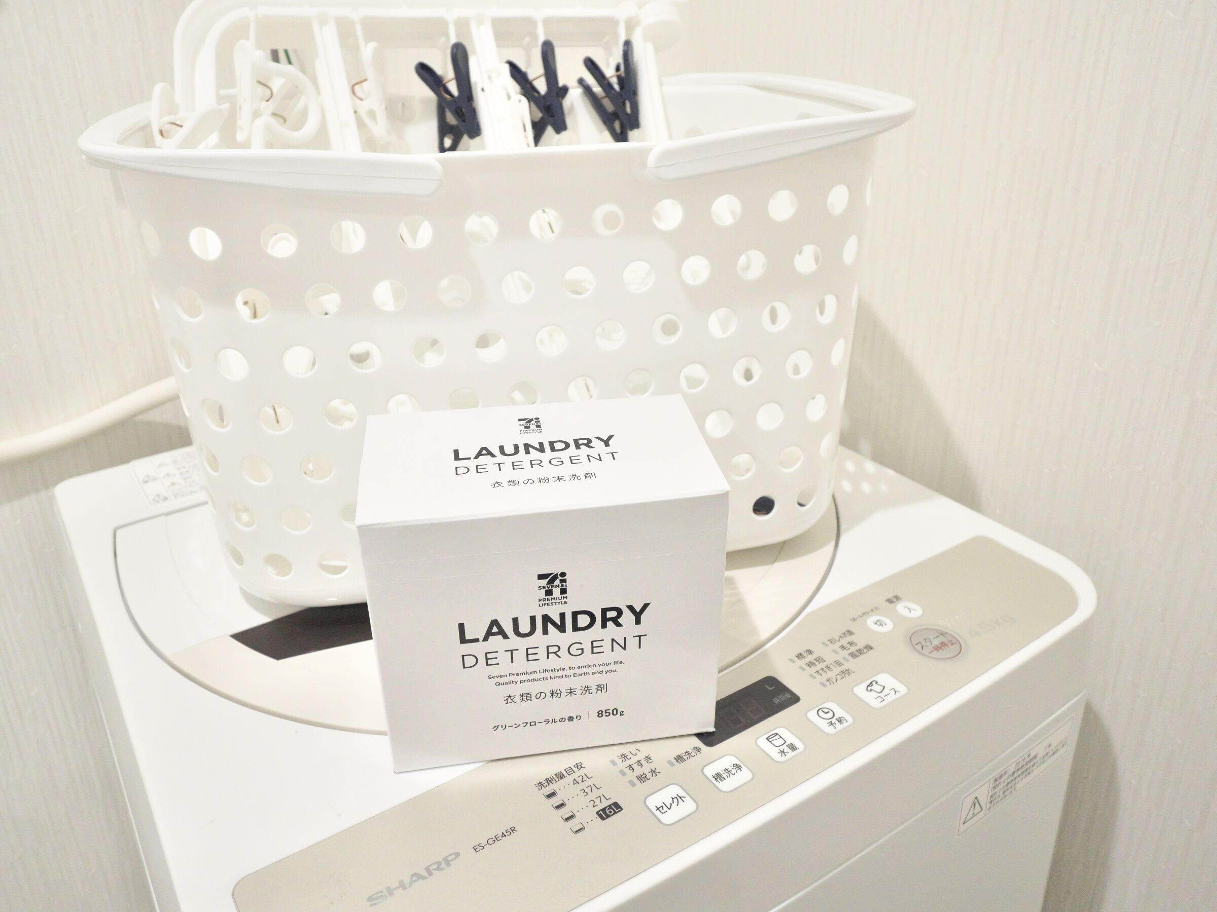 A white laundry detergent box on a washing machine with a white perforated basket containing clothespins.