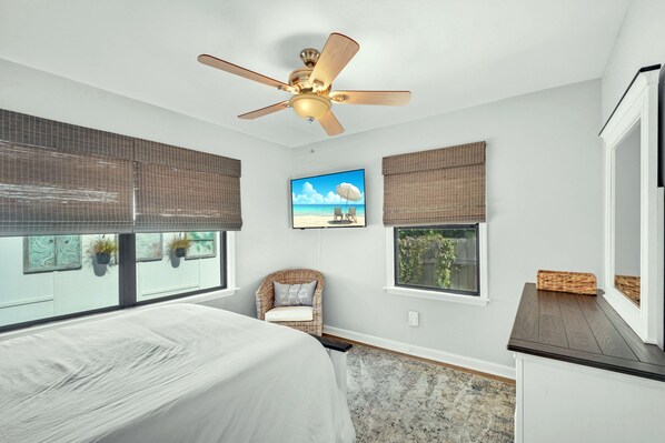 A bedroom with a bed, a ceiling fan, a TV, and large windows with blinds.