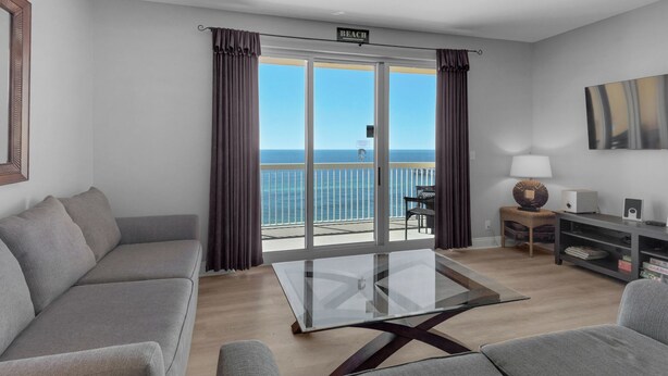 A modern living room with a glass table, grey sofas, a TV, and a balcony with a view of the sea. - Calypso Resort by BBG (Panama City Beach)