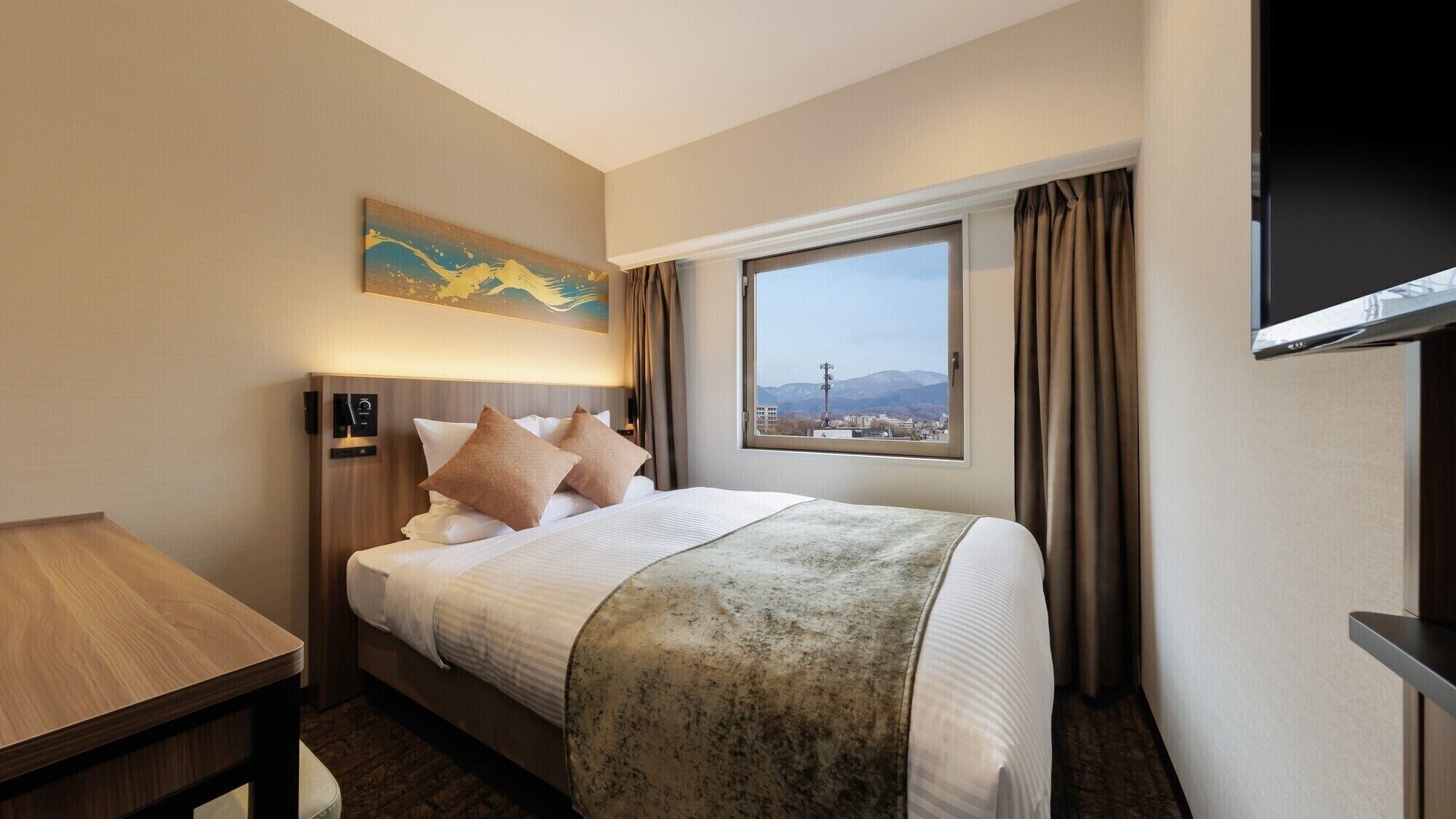 A hotel room with a bed, a desk, a painting, a window with a view, and a TV.