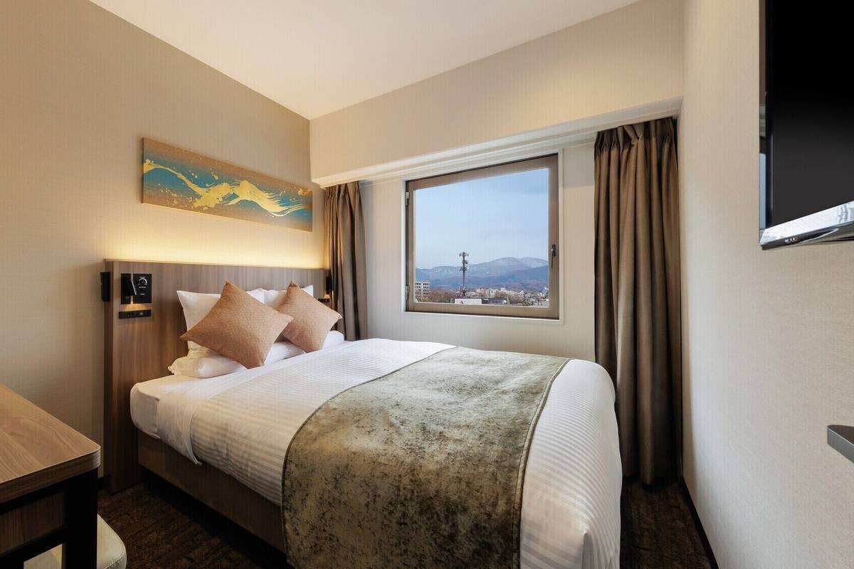 A hotel room with a bed, a desk, a painting, a window with a view, and a TV.