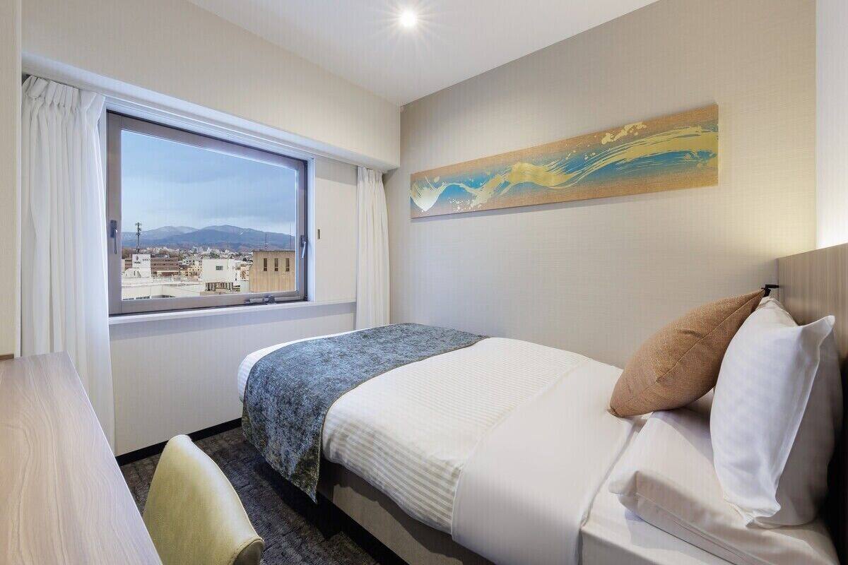 A hotel room with a bed, a desk, a chair, a window with a view of buildings, and a painting on the wall.