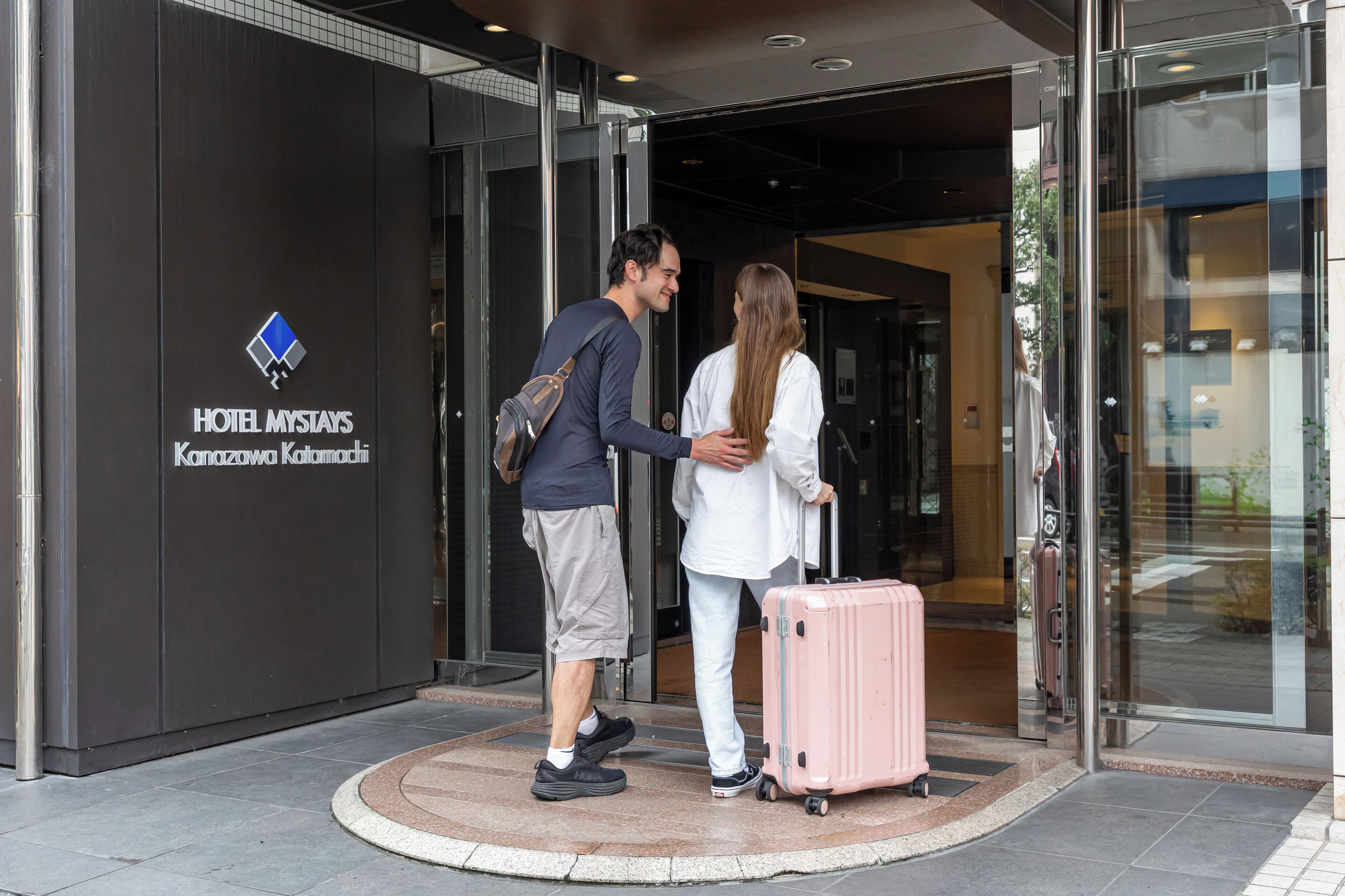 Egy férfi és egy nő áll egy szálloda bejáratánál, a bejárat felett egy tábla látható, amelyen a következő felirat szerepel: „HOTEL MYSTAYS”.
