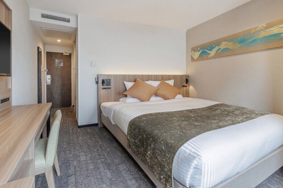 A hotel room with a bed, a desk, and a chair. There is a painting on the wall and a door leading to another room.