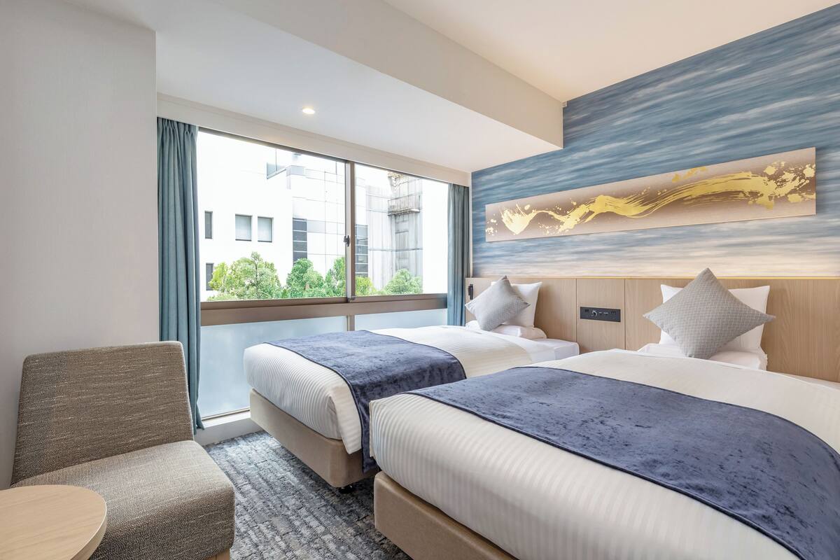 A hotel room with two beds, a small table, and a view of greenery outside.