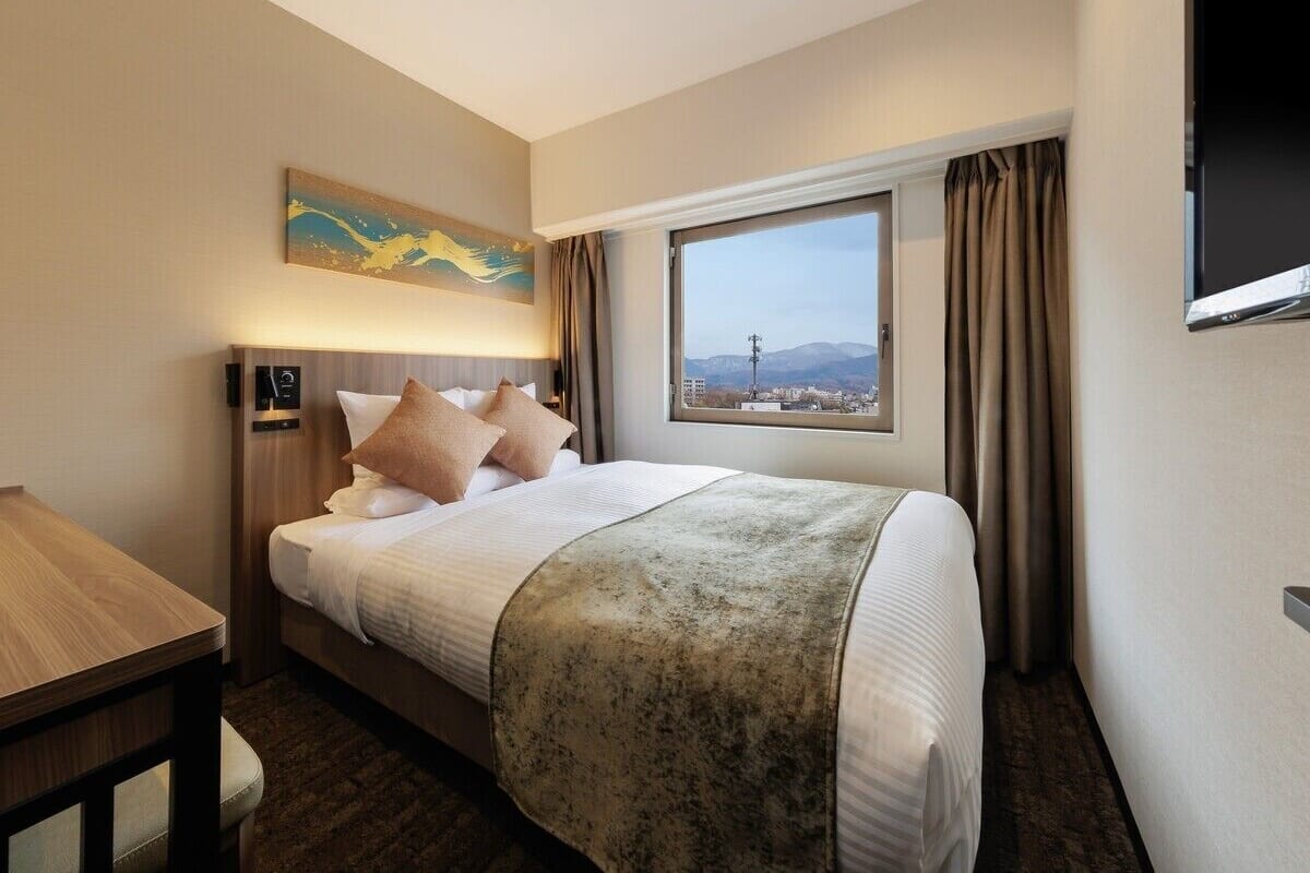 A hotel room with a bed, a bedside table, a painting on the wall, a window with curtains, and a view of the cityscape.