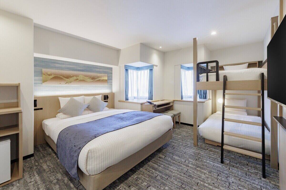 A modern hotel room with a large bed, a desk, a bunk bed, and a large window with curtains.