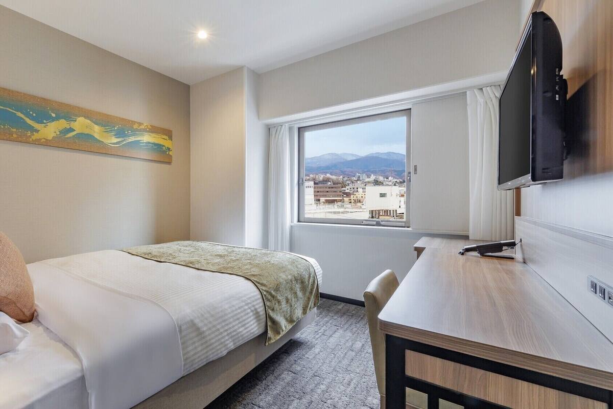 A hotel room with a bed, a desk, a television, a window with a city view, and a painting on the wall.
