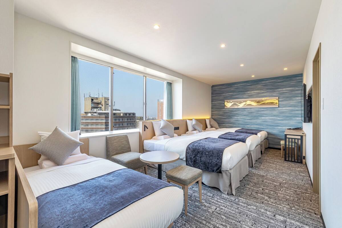 A hotel room with three beds, a small round table, and a city view.